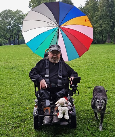 Andreas Vega steht auf einer Wiese neben seiner Hündin Berta. Sein Rollstuhl wird von einem mehrfarbigen Schirm vor der Witterung geschützt.