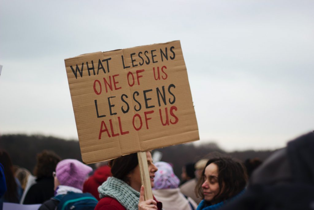 Menschen halten auf einer Demonstration ein Schild in die Höhe, darauf steht "What lessens one of us lessens all of us". Auf Deutsch "Was einen von uns benachteiligt, benachteiligt uns alle"